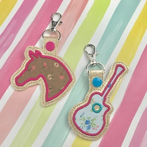 以下が含まれることがあります： 花柄の生地のデザインが施されたキーホルダーが2つ。1つは馬の頭、もう1つはギター。両方のキーホルダーはピンクのステッチと金色の金属リングが付いています。