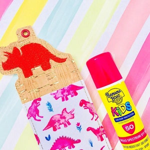 Puede incluir: Un llavero de tela con estampado de dinosaurios rosa y blanco con un dinosaurio rojo en la parte superior. Un stick de protector solar rosa con el texto "Banana Boat Kids SPF 50" está al lado del llavero.