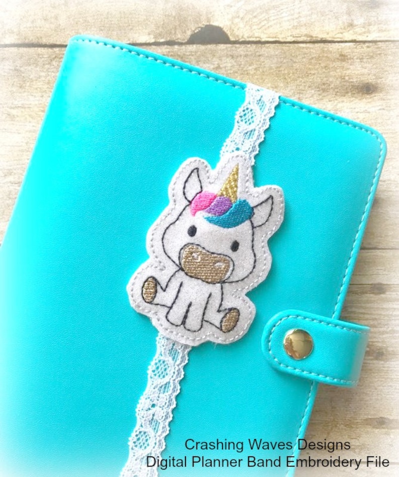May include: A white unicorn planner band with a gold horn, pink and blue mane, and a white lace trim. The band is attached to a turquoise planner.