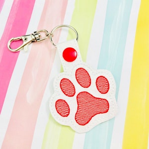 May include: White paw print keychain with a red embroidered paw print and a red button. The keychain has a silver metal clasp.