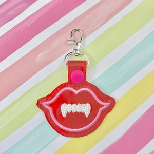 May include: Red fabric keychain with a vampire mouth design. The mouth has white fangs and a pink lip. The keychain has a silver metal ring.