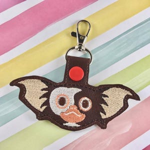 May include: A brown embroidered keychain featuring a Gremlin character design. The keychain has a silver clasp and a brown leather backing with a red button. The Gremlin has tan ears, white eyes, and an orange mouth. The background is a colorful striped pattern.
