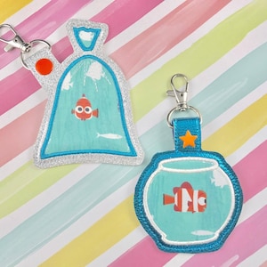 May include: Two keychains featuring a cartoon fish in a plastic bag and a fishbowl. The plastic bag keychain is silver with a blue and white design. The fishbowl keychain is blue with a white and blue design.