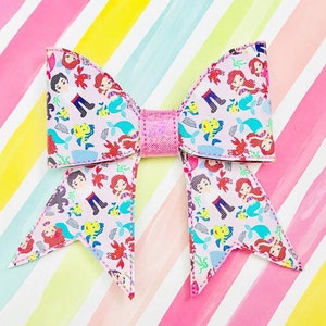 May include: A pink bow with a mermaid and Prince Eric print. The bow is made of fabric and has a glittery centre.