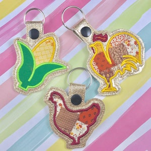 May include: Three gold keychains with different embroidered designs. One keychain features a green and yellow corn cob, another features a red and brown rooster, and the last keychain features a red and brown hen.