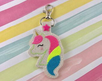 May include: A white unicorn keyring with a pink, yellow and blue mane and tail. The unicorn has a gold horn and a pink star on its head. The keyring is attached to a silver metal key ring.