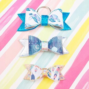 May include: Three colorful fabric bows with different patterns. The top bow is blue with a white background and a sea turtle design. The middle bow is blue and silver with a white background and a jellyfish design. The bottom bow is white with a gold background and a feather design.