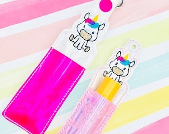 Puede incluir: Dos porta-gel de manos de plástico transparente con forma de unicornio. Un porta-gel es rosa y el otro es iridiscente. Ambos porta-geles tienen un unicornio blanco con una crin y un cuerno arcoíris bordados en la parte delantera. El porta-gel rosa tiene una cadena de llaves plateada.