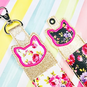 May include: Two gold glitter lip balm holders with floral fabric and pink cat-shaped embroidery. One holder has a white heart and a silver keychain.