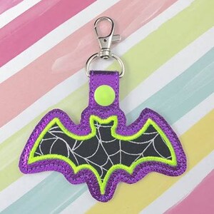 May include: Purple glitter bat keychain with a black spiderweb design and a lime green outline. The keychain has a silver metal clip.