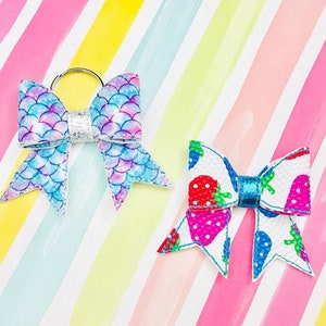 May include: Two fabric hair bows with a key ring attached to one bow. The bow on the left is blue and purple with a mermaid scale pattern. The bow on the right is white with a strawberry pattern and a turquoise blue center.