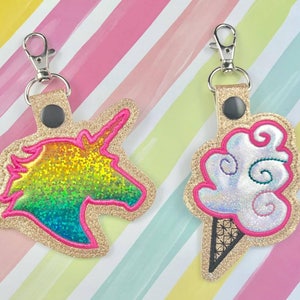 May include: Two keychains with gold glitter fabric and pink stitching. One keychain features a rainbow glitter unicorn head and the other features a white glitter ice cream cone with a swirl of blue and green.