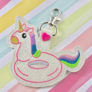 Op de afbeelding: Een witte glitter sleutelhanger met een eenhoorn die op een opblaasbaar ringetje rijdt. De eenhoorn heeft een roze, paarse en groene manen en staart. De ring is roze en heeft een zilveren binnenring. De sleutelhanger heeft een roze hartje.
