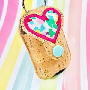 May include: A cork and fabric hand sanitizer holder with a pink heart-shaped flap. The heart is decorated with a floral pattern and a pink border. The holder has a light blue button and a silver key ring.