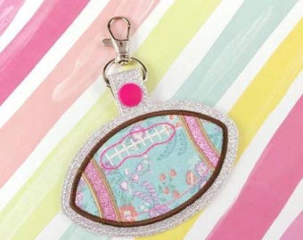 Puede incluir: Un llavero de fútbol de color plateado con purpurina, con un botón rosa y un clip plateado. El balón de fútbol está decorado con un patrón floral azul y rosa.