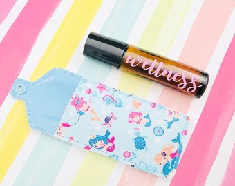 Puede incluir: Una bolsa de tela azul y blanca con un diseño de sirena y una botella de vidrio marrón con rodillo y tapa negra. La botella tiene una etiqueta rosa que dice "wellness".
