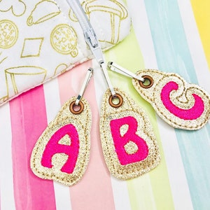 Puede incluir: Tres tiradores de cremallera dorados con letras A, B y C de fieltro rosa. Los tiradores de cremallera están sobre un fondo a rayas.