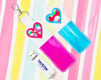 Puede incluir: Dos llaveros con diseños en forma de corazón y una unidad USB. Un llavero es rosa intenso, el otro es azul. Los corazones tienen patrones coloridos tipo confeti. La unidad USB es blanca con el texto "brother at your side."