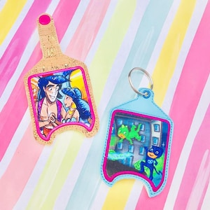 May include: Two keychains with cartoon images. One keychain is tan with a pink border and features Aladdin and Jasmine. The other is light blue with a pink border and features characters from PJ Masks. Both have a metal keyring.