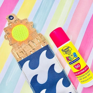 May include: A blue and white wave-patterned fabric pouch with a cork and orange sun design. The pouch holds a pink and yellow sunscreen stick labeled "Banana Boat Kids SPF 50".