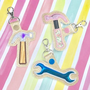 May include: Three gold glitter keychains with different tool designs. One keychain features a pink hammer with a floral pattern, another has a blue wrench, and the last one has a purple hammer with a wood-grain handle.