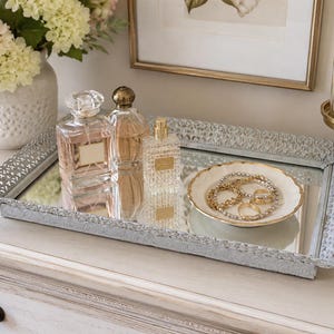 May include: A rectangular mirrored vanity tray with an ornate, light gray metal filigree border. The tray holds three glass perfume bottles of varying shapes and a small, round white ceramic dish with a gold rim containing gold-toned jewelry.