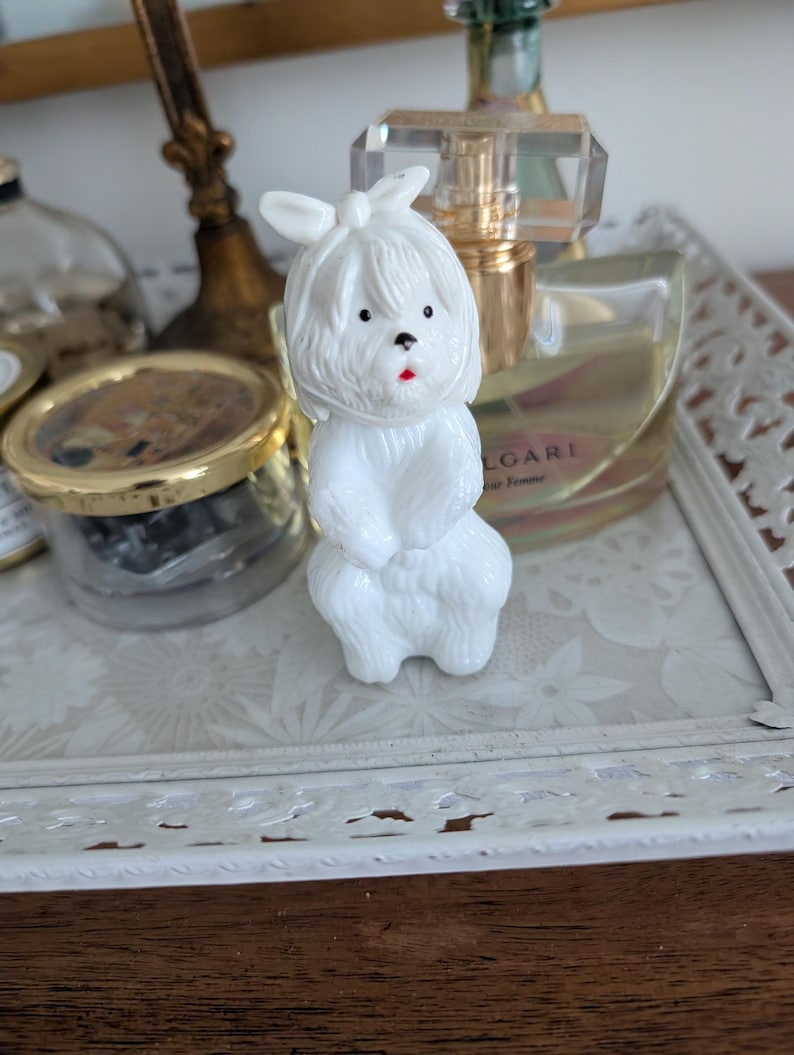 K&ouml;nnte beinhalten: Kleine, wei&szlig;e Hundefigur mit Schleife auf dem Kopf. Der Hund hat schwarze Augen und einen roten Mund. Die Figur steht auf einem wei&szlig;en Tablett mit floralem Muster. Weitere Gegenst&auml;nde sind im Hintergrund zu sehen.