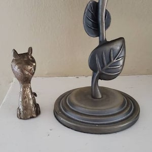 Puede incluir: Una pequeña figurita de animal estilizada de color bronce se encuentra sobre una superficie blanca delante de una lámpara con una pantalla de cristal de colores y una base con diseño de hojas.