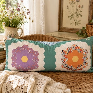 Op de afbeelding: Een rechthoekig sierkussen met een groenblauwe achtergrond en twee grote bloemmotieven gemaakt van zeshoekig patchwork. De linkerbloem heeft een geel hart met paarse bloemblaadjes, de rechterbloem heeft een geel hart met oranje bloemblaadjes.