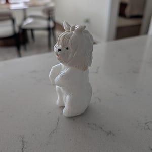 K&ouml;nnte beinhalten: Kleine, wei&szlig;e Hundefigur mit einer Schleife auf dem Kopf. Der Hund steht auf seinen Hinterbeinen, die Vorderpfoten sind zusammengelegt. Er hat schwarze Augen und eine rote Nase. Die Figur besteht aus einem glatten, gl&auml;nzenden Material.