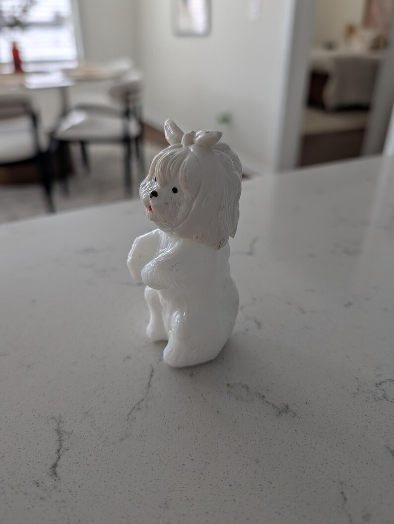 K&ouml;nnte beinhalten: Kleine, wei&szlig;e Hundefigur mit einer Schleife auf dem Kopf. Der Hund steht auf seinen Hinterbeinen, die Vorderpfoten sind zusammengelegt. Er hat schwarze Augen und eine rote Nase. Die Figur besteht aus einem glatten, gl&auml;nzenden Material.