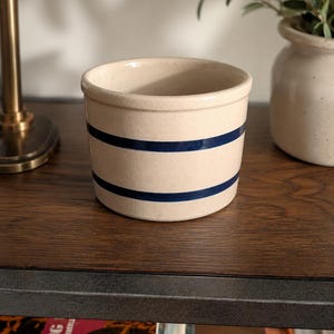 Könnte beinhalten: Ein cremefarbener Keramiktopf mit zwei marineblauen Streifen. Der kleine, runde Behälter steht auf einer dunklen Holzoberfläche. Ein dekoratives Stück im Vintage-Stil, wahrscheinlich zur Aufbewahrung oder Präsentation.