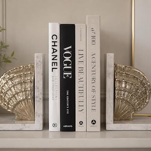 May include: Decorative seashell bookends in gold, resting on white marble bases, flank a collection of books. The books include titles like "CHANEL," "VOGUE," and "A Century of Style." The bookends and books are arranged on a white surface.