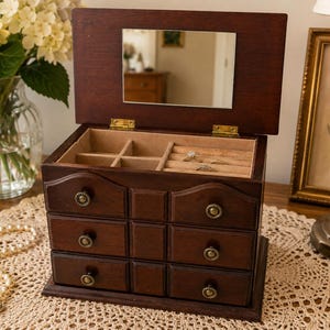 May include: A dark brown wooden jewelry box featuring a hinged lid with an interior rectangular mirror, three tiers of drawers with small round brass-toned knobs, and a top compartment lined with beige fabric containing ring slots and dividers.