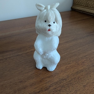 K&ouml;nnte beinhalten: Eine wei&szlig;e Hundefigur mit Schleife auf dem Kopf und rotem Mund. Der Hund hat schwarze Augen und ein flauschiges Aussehen. Die Figur besteht aus einem glatten Material und steht auf einer Holzoberfl&auml;che.