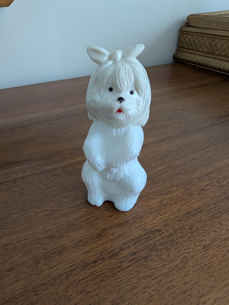 K&ouml;nnte beinhalten: Eine wei&szlig;e Hundefigur mit Schleife auf dem Kopf und rotem Mund. Der Hund hat schwarze Augen und ein flauschiges Aussehen. Die Figur besteht aus einem glatten Material und steht auf einer Holzoberfl&auml;che.