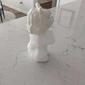 K&ouml;nnte beinhalten: Eine wei&szlig;e Keramik-Hundefigur mit strukturiertem Fell und aufrechten Ohren. Der Hund sitzt mit zusammengelegten Vorderpfoten. Die Figur steht auf einer hellen Arbeitsplatte.
