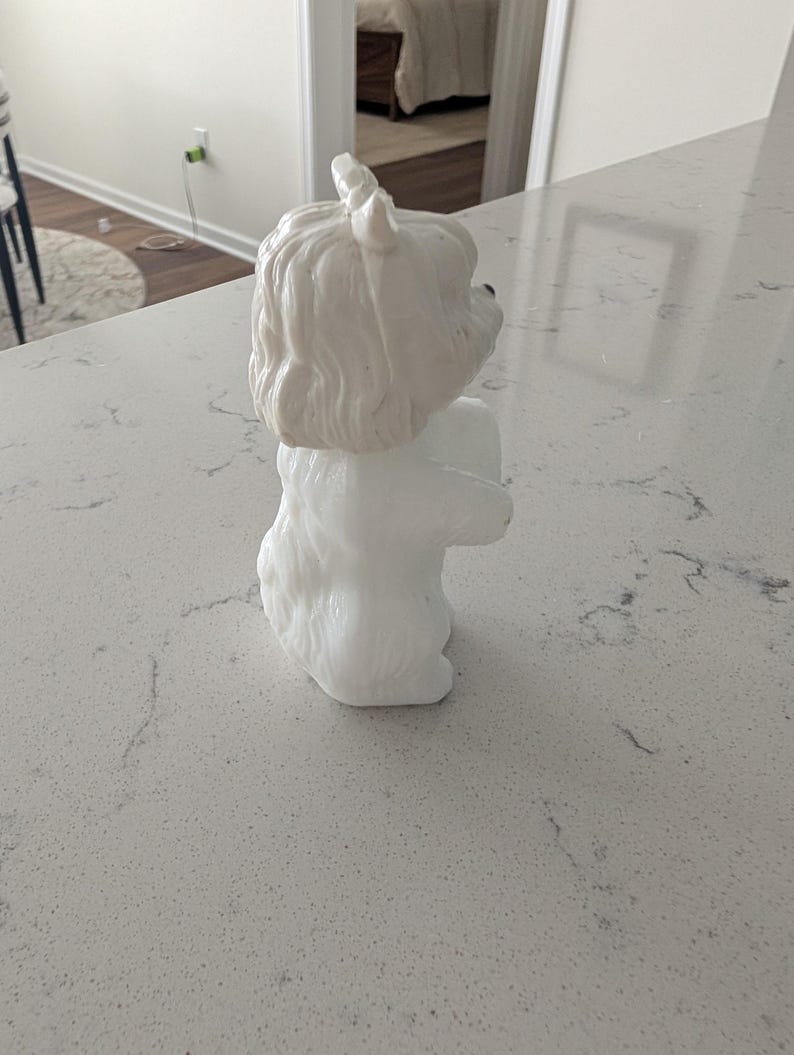 K&ouml;nnte beinhalten: Eine wei&szlig;e Keramik-Hundefigur mit strukturiertem Fell und aufrechten Ohren. Der Hund sitzt mit zusammengelegten Vorderpfoten. Die Figur steht auf einer hellen Arbeitsplatte.