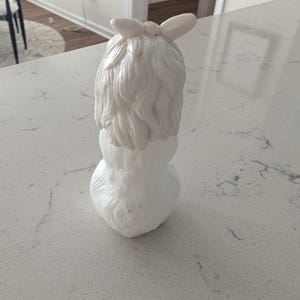 K&ouml;nnte beinhalten: Eine wei&szlig;e Keramikfigur einer Frau mit einer Schleife im Haar. Die Figur hat eine detaillierte Frisur und einen abgerundeten Sockel. Das Gesamtdesign ist glatt und elegant, mit gl&auml;nzendem Finish.