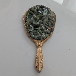 May include: A vintage hand mirror with a gold-colored ornate handle and frame. The mirror's surface is covered with small, green, irregularly shaped stones. The handle has a detailed floral design.