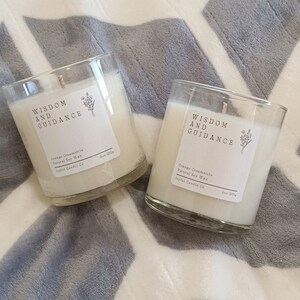 Puede incluir: Dos velas de vidrio transparente con cera blanca y una etiqueta que dice "WISDOM AND GUIDANCE". La etiqueta también dice "Orange Creamsicle Natural Soy Wax". Las velas están sobre una superficie con estampado gris y blanco.