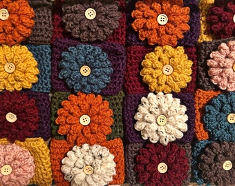 May include: A crocheted blanket with a patchwork design featuring various colors and textures. Each square features a crocheted flower with a wooden button in the center. The colors include burgundy, mustard yellow, teal, cream, and brown.