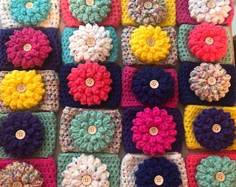 May include: A collection of crocheted cup cozies with various colors and patterns. Each cozy features a crocheted flower with a button center.