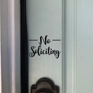 May include: A white door with a black door frame and a black door handle. A black vinyl sticker on the door reads "No Soliciting".