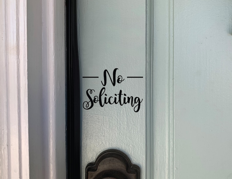 May include: A white door with a black door frame and a black door handle. A black vinyl sticker on the door reads "No Soliciting".