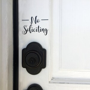 May include: A white door with a black door knob and a black vinyl sticker that says "No Soliciting" in cursive.