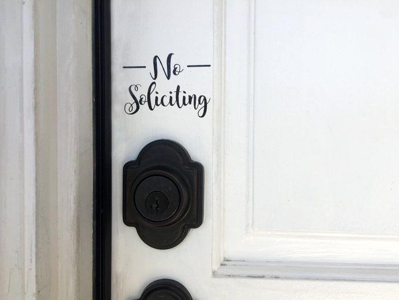 May include: A white door with a black door knob and a black vinyl sticker that says "No Soliciting" in cursive.
