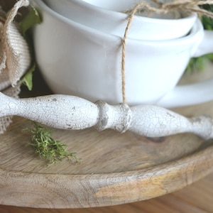 May include: A rustic wooden tray holds two white ceramic teacups tied with twine, a distressed white decorative spindle, and a burlap flower. The tray has a natural wood grain finish, and the overall aesthetic is farmhouse style.