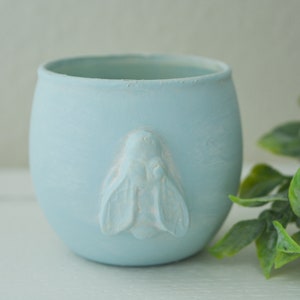 Puede incluir: Una maceta de cerámica azul claro con un diseño de abeja. La maceta redonda y pequeña tiene un acabado mate y una aplicación de abeja en relieve en la parte delantera. Follaje artificial verde y amarillo en el fondo.