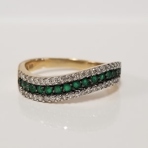 Op de afbeelding: Een gouden ring met een golvend ontwerp. De ring heeft drie rijen edelstenen: een centrale rij groene smaragden, geflankeerd door rijen kleine, heldere diamanten. De ring staat tegen een witte achtergrond.